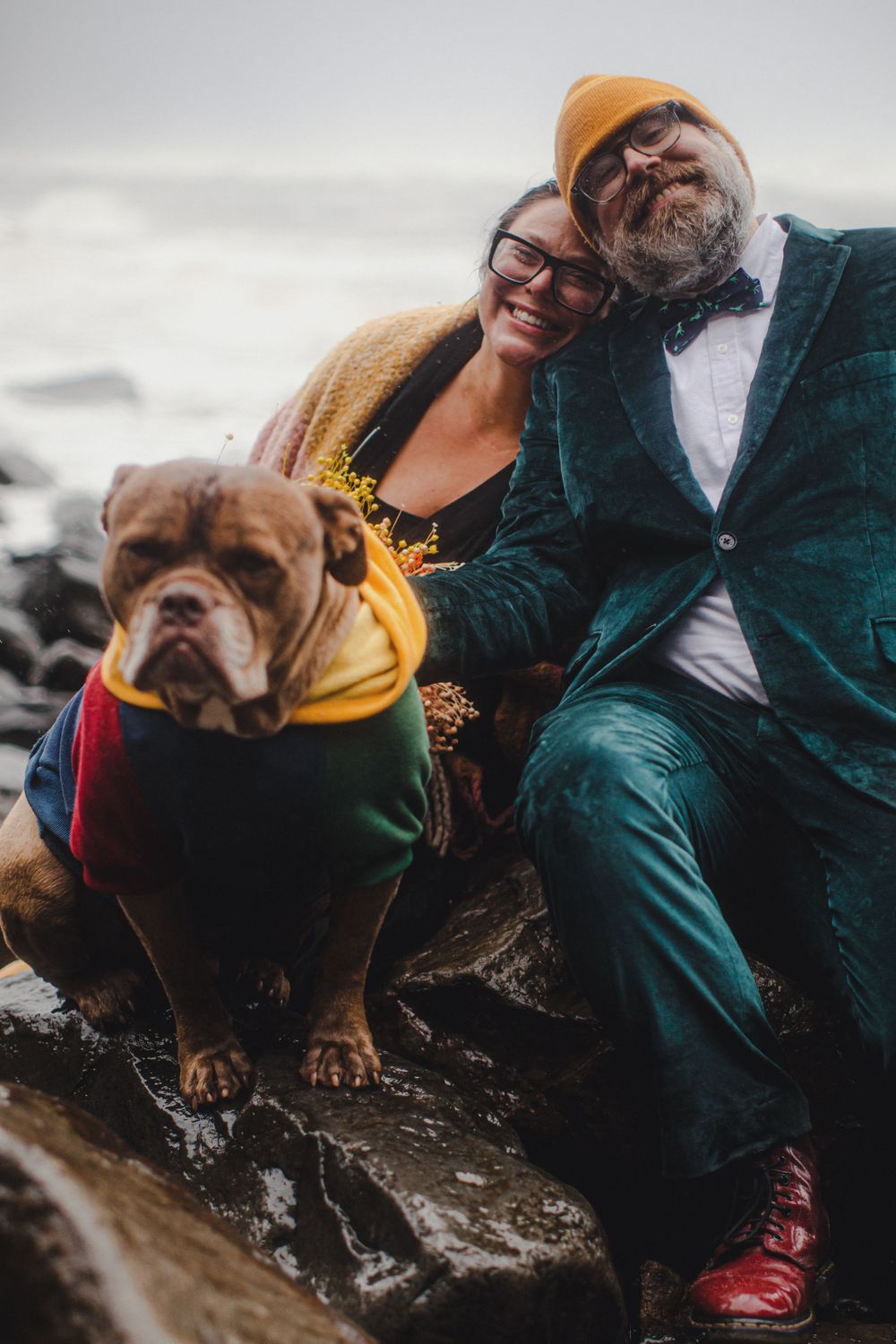 A couple and their English Bulldog in formal wear pose together on rocky beach terrain.