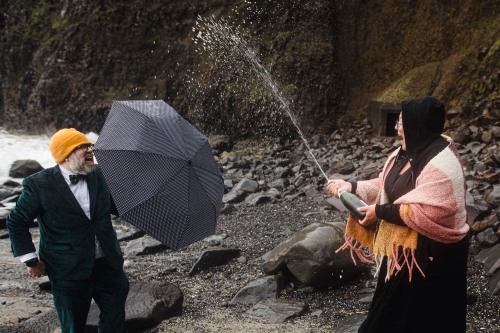 Two elegantly dressed figures with umbrellas stand on rocky terrain near a waterfall.
