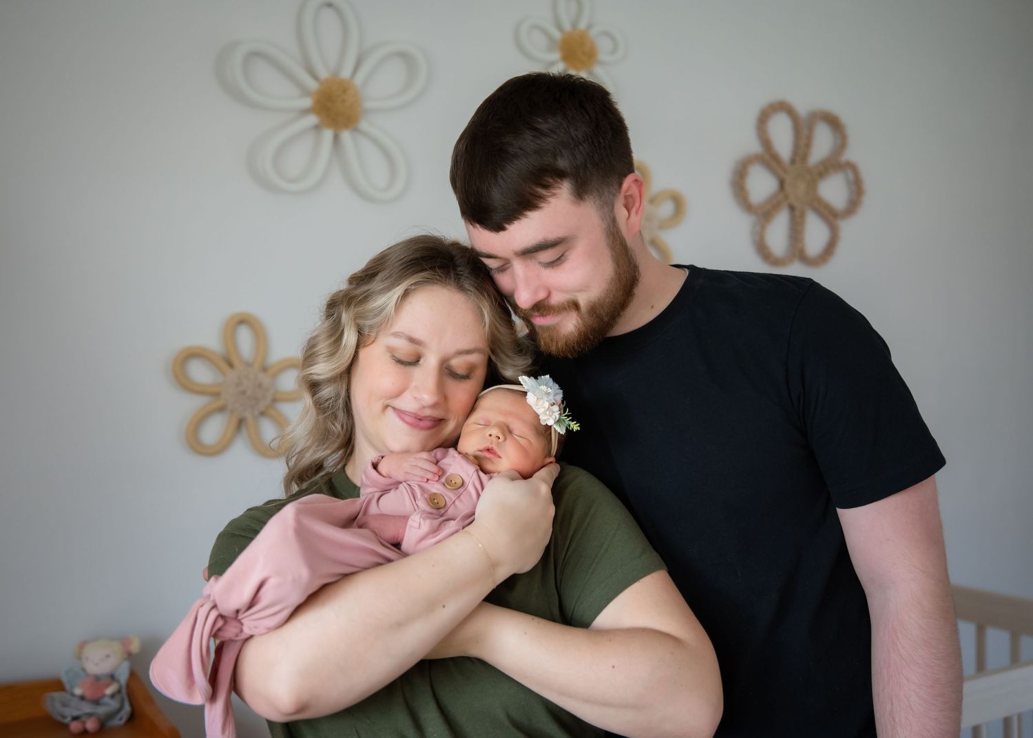 Parents hold their sleeping newborn baby while sitting against a wall decorated with floral designs.