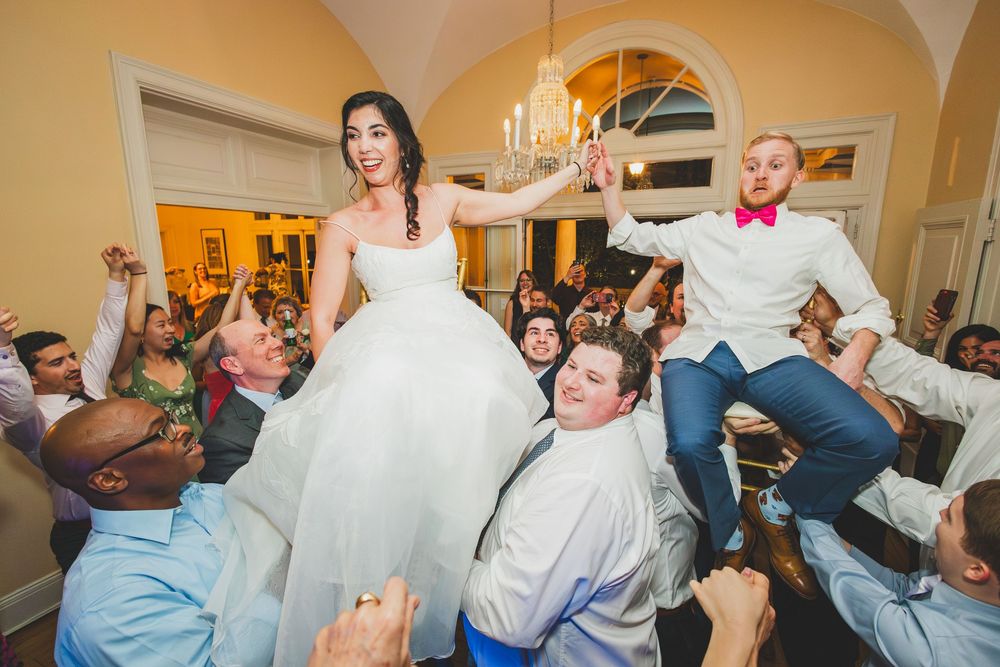 Wedding reception celebration with guests lifting the newlyweds on chairs during traditional hora dance at indoor venue.