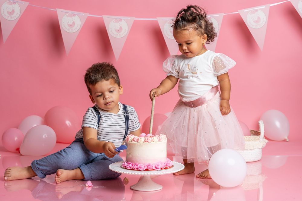 Baby in white dress crawling towards pink birthday cake with balloon decorations in pink studio setting.