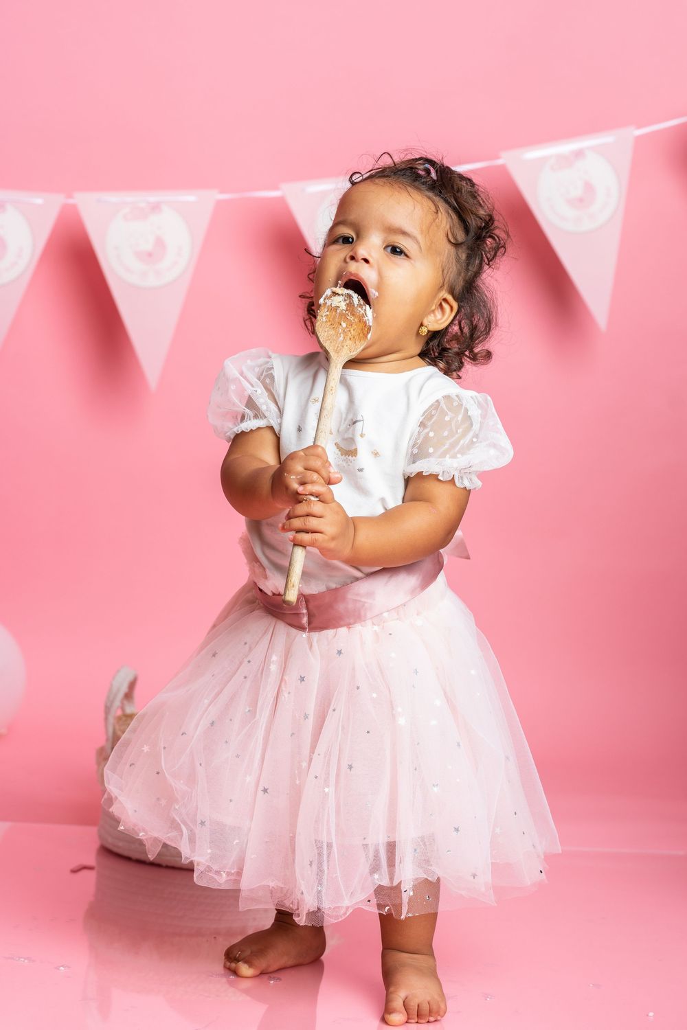 Toddler in white lace dress and tulle skirt enjoying cake on a stick against pink backdrop with banner decorations.