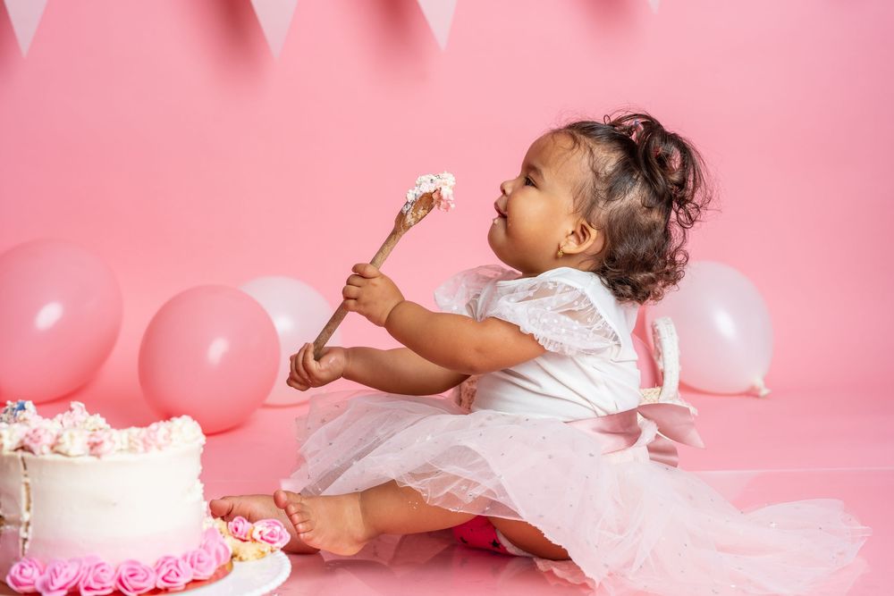 Toddler in white dress sitting on pink backdrop with birthday cake and balloons enjoying cake with a spoon.