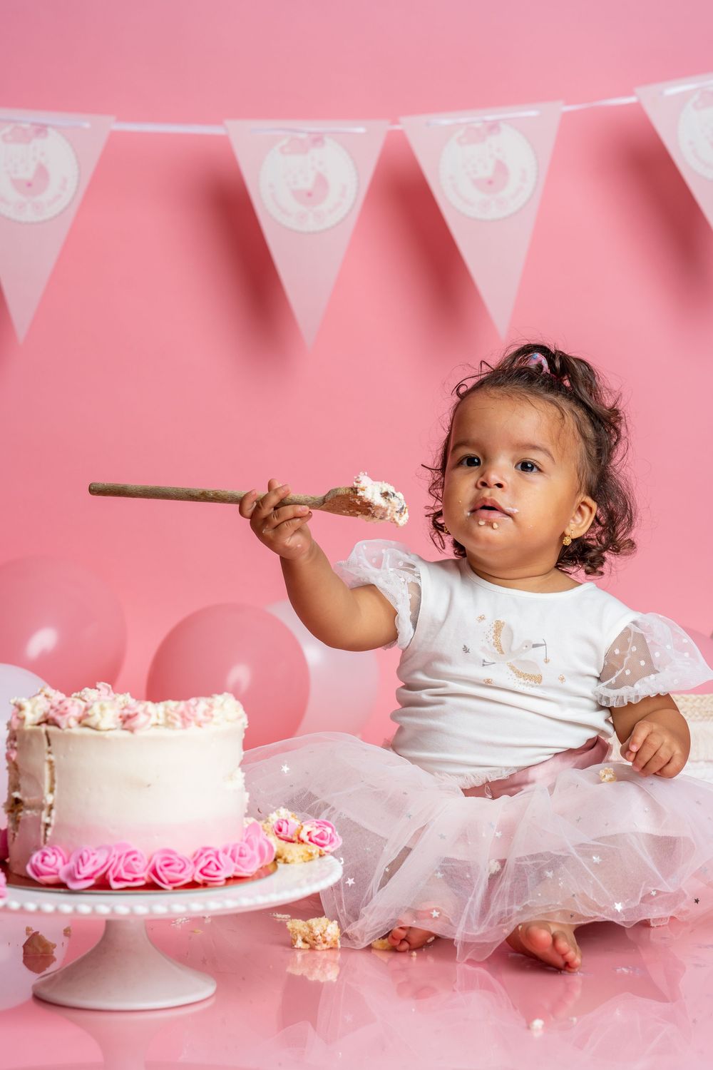 Small child in tutu dress samples cake during photo shoot with pink and white birthday decorations.
