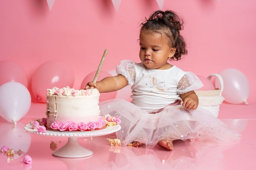 Baby sits near decorated birthday cake in tulle dress during pink-themed cake smash photoshoot.