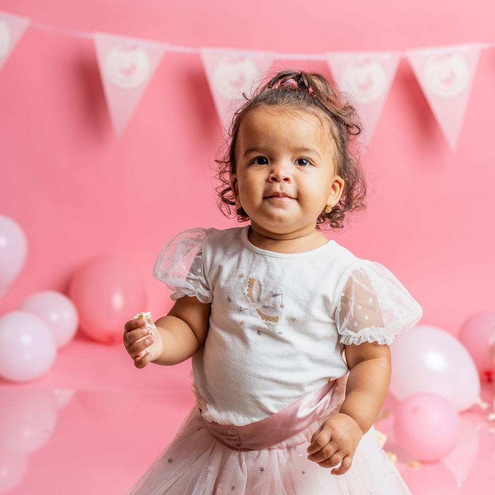 Baby in white lace dress sitting with birthday decorations and pink balloons.