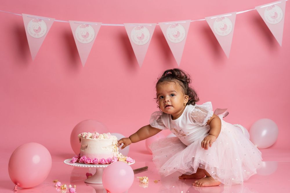 Baby reaching for pink birthday cake against pink backdrop with birthday banners and decorations.