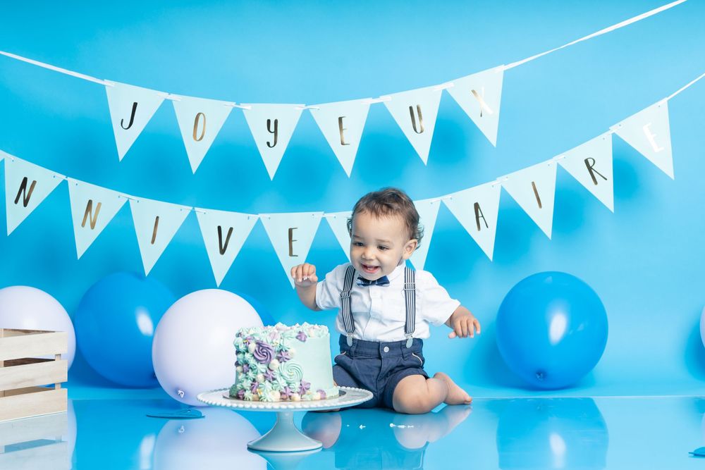 A festive first birthday party setup with white bunting and blue balloons against a bright turquoise background.