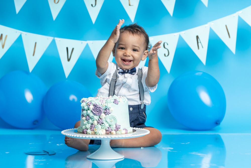 A festive cake smash photo setup with white bunting, blue balloons and a decorated white cake on a stand.