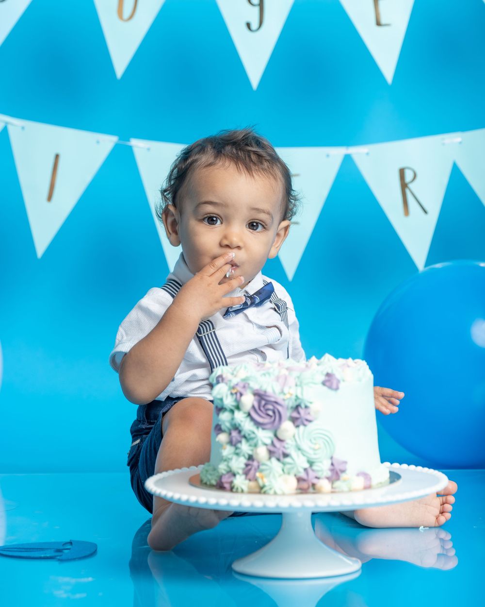 A birthday cake decorated in purple and teal sits on a white pedestal against a bright blue backdrop with white banner flags.