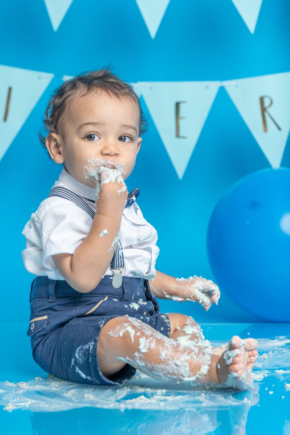 A messy cake smash photo session with white cake pieces scattered on a blue background with white banner decorations.