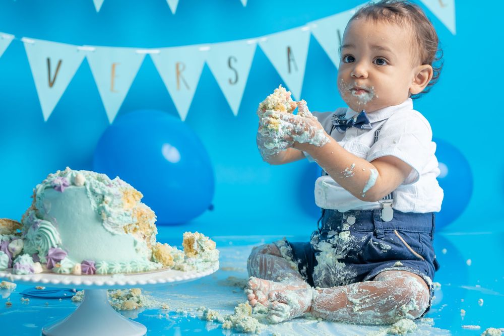 A cake smash photo session with baby in formal wear surrounded by blue balloons and white decorative banners.