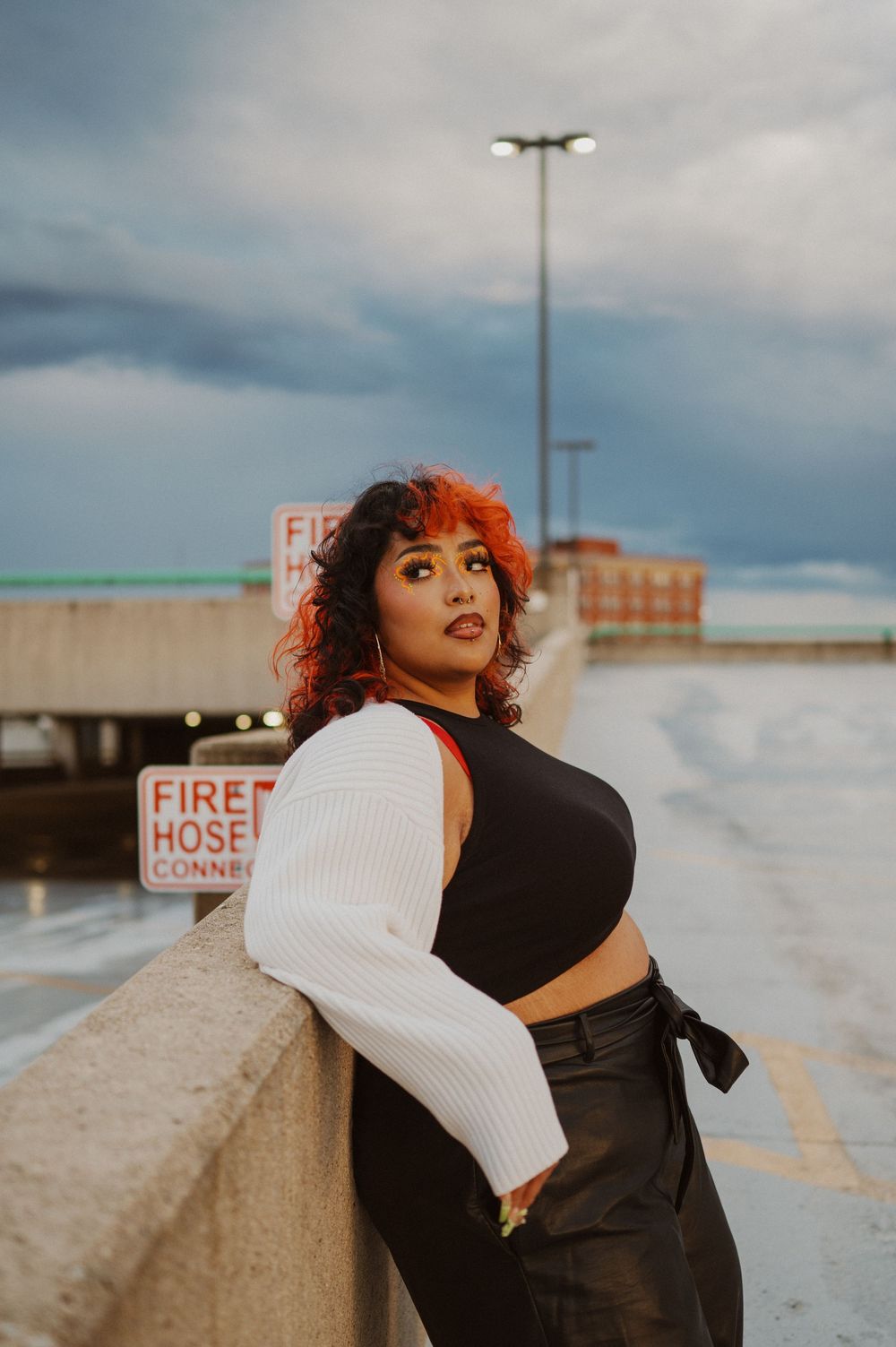 A fashionable model in black and white streetwear poses against an industrial backdrop on an overcast day.