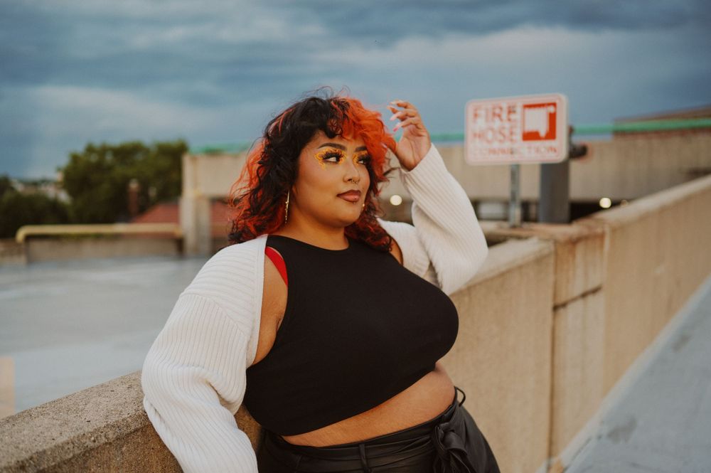 Person wearing a white cropped sweater and black top poses casually on a rooftop with basketball hoop in background.