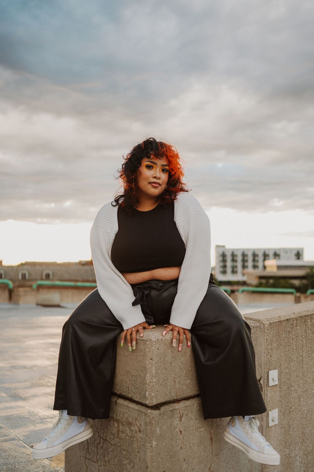 A person in a black dress and white sleeves sits on a stone wall against a cloudy sky while looking at the camera.