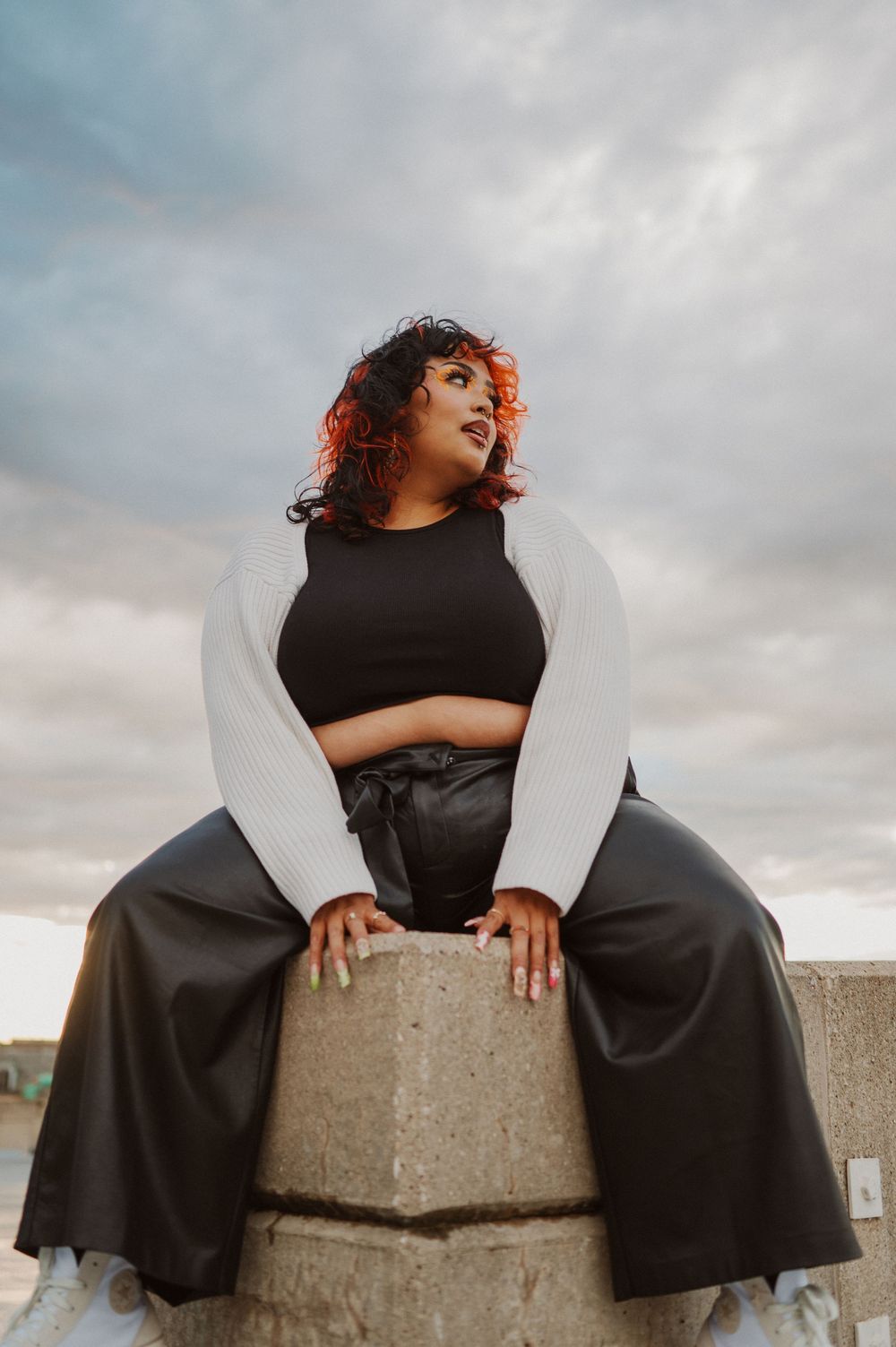 A person in black clothing sits gracefully on a concrete ledge against moody cloudy skies during a dramatic photoshoot.