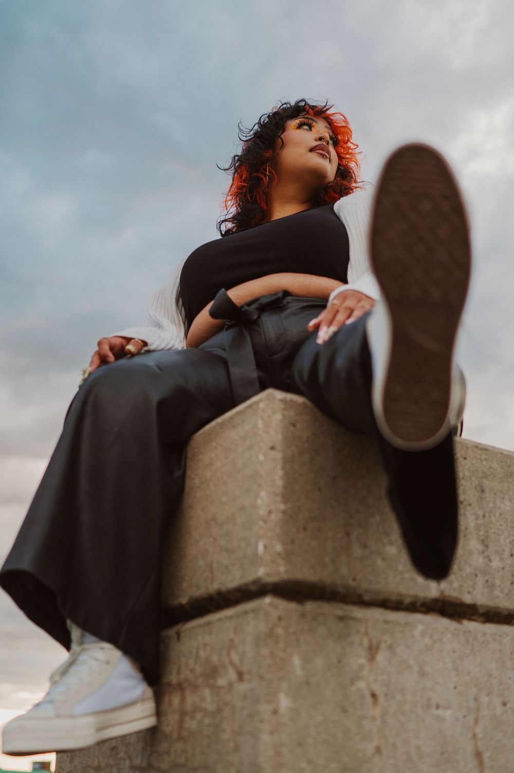 Person in black outfit lounges on concrete structure against cloudy sky, showing sneaker sole in artistic low angle shot.