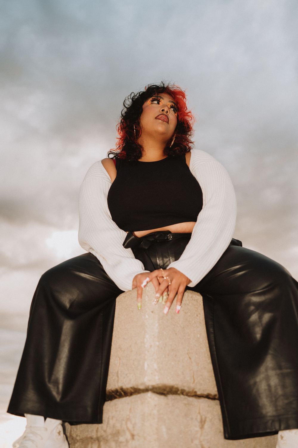 A person wearing a black crop top and leather pants poses on a concrete block in dramatic lighting against cloudy skies.
