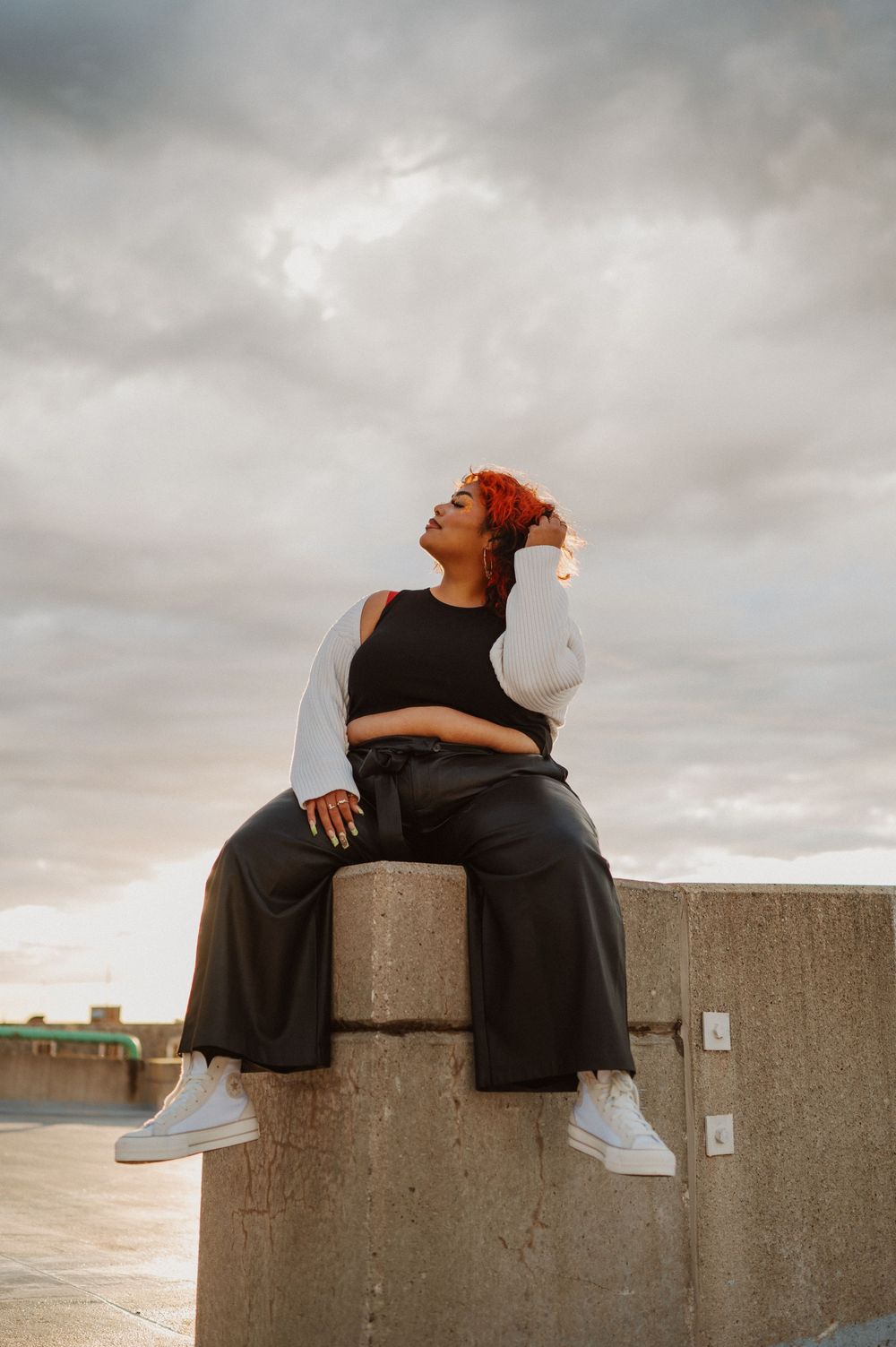 Someone in casual wear sits thoughtfully on a concrete wall against a cloudy sky at sunset.