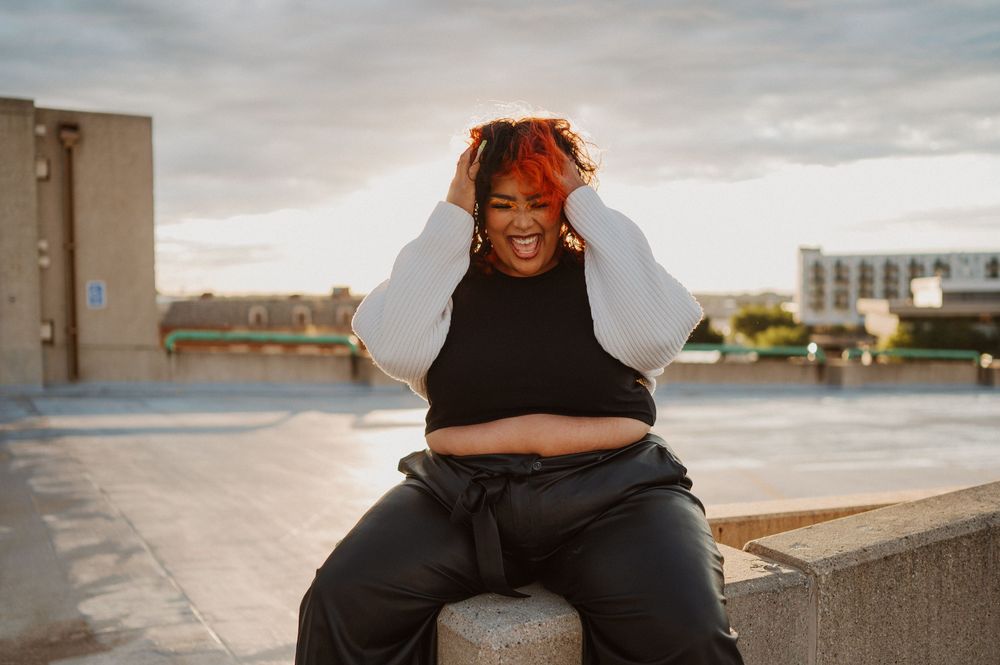 A person with bright orange hair wearing a black crop top sits outdoors on a concrete ledge on a cloudy day.