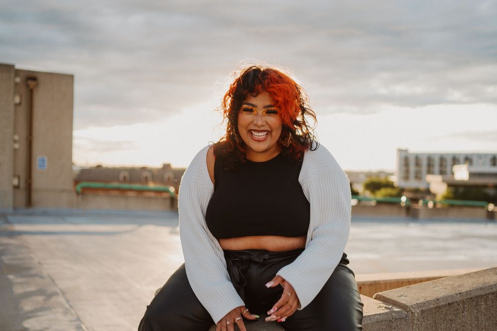 A person in a gray sweater and black crop top sits smiling on an urban rooftop during sunset.
