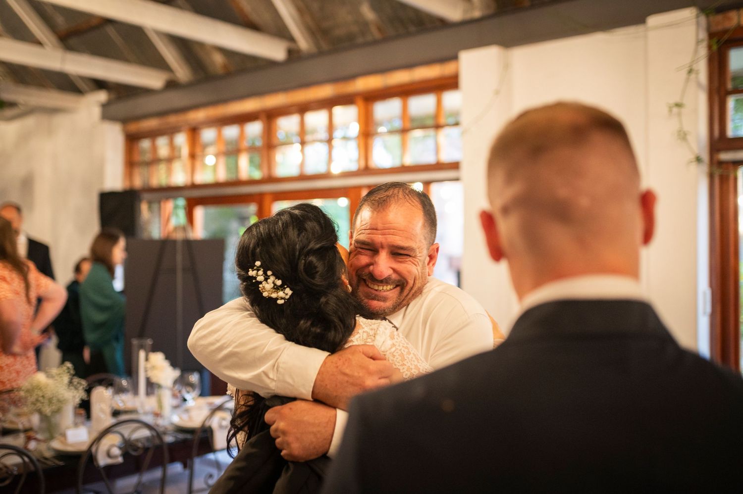 Wedding celebration scene showing guests dancing and embracing in a rustic indoor venue with natural lighting.