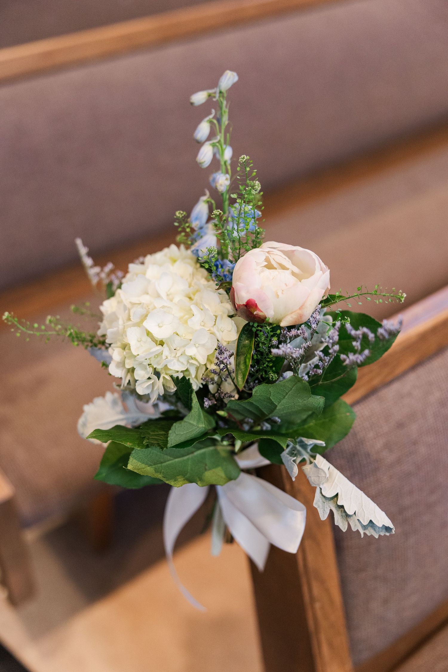 Delicate floral pew decoration with white hydrangea, blush rose, and greenery tied with ribbon for a church wedding ceremony.