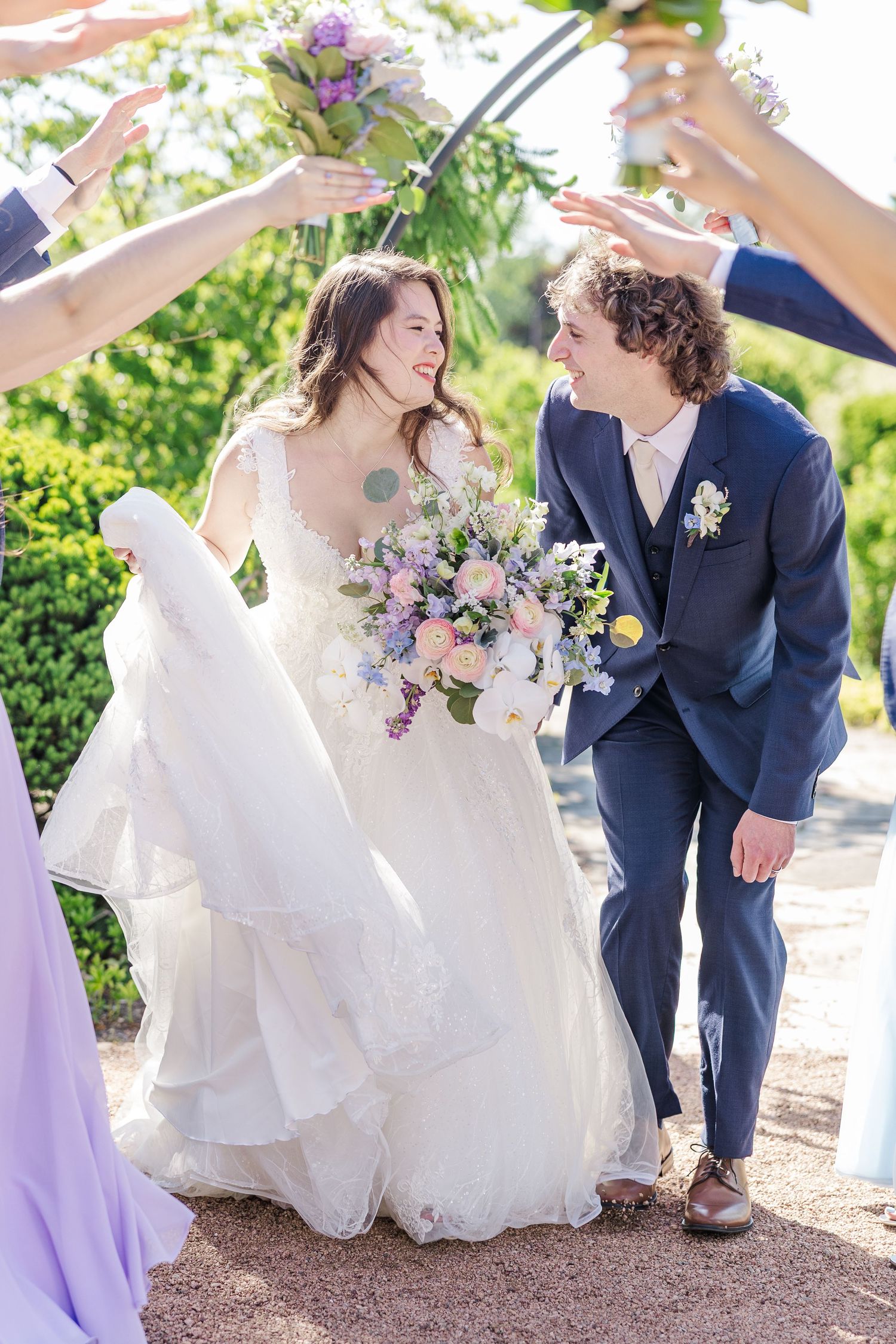 A joyful wedding celebration outdoors with a bride in a flowing white gown and groom in navy suit under floral decorations.