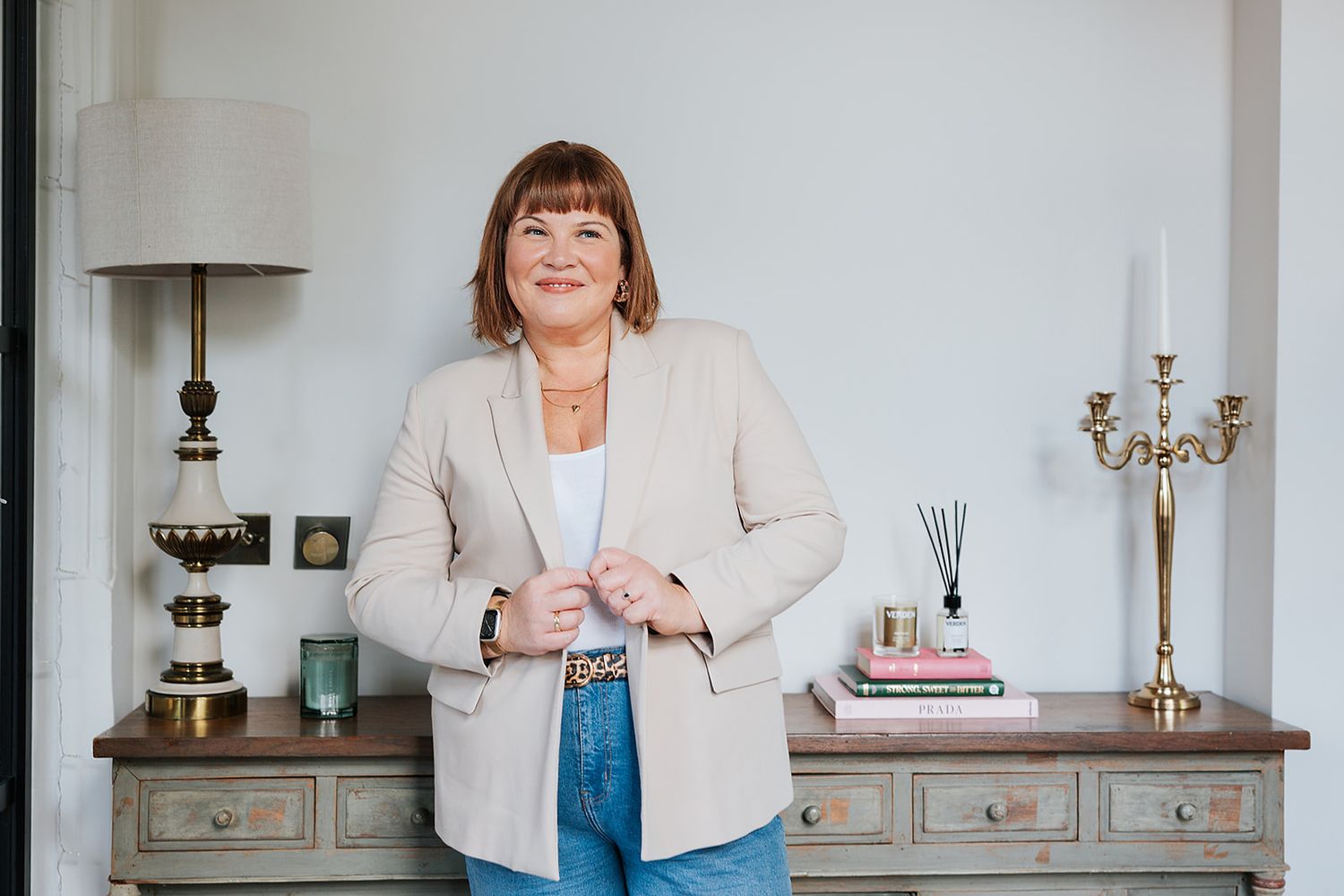 Professional wearing a beige blazer and jeans stands in front of a vintage console table with home decor accents.