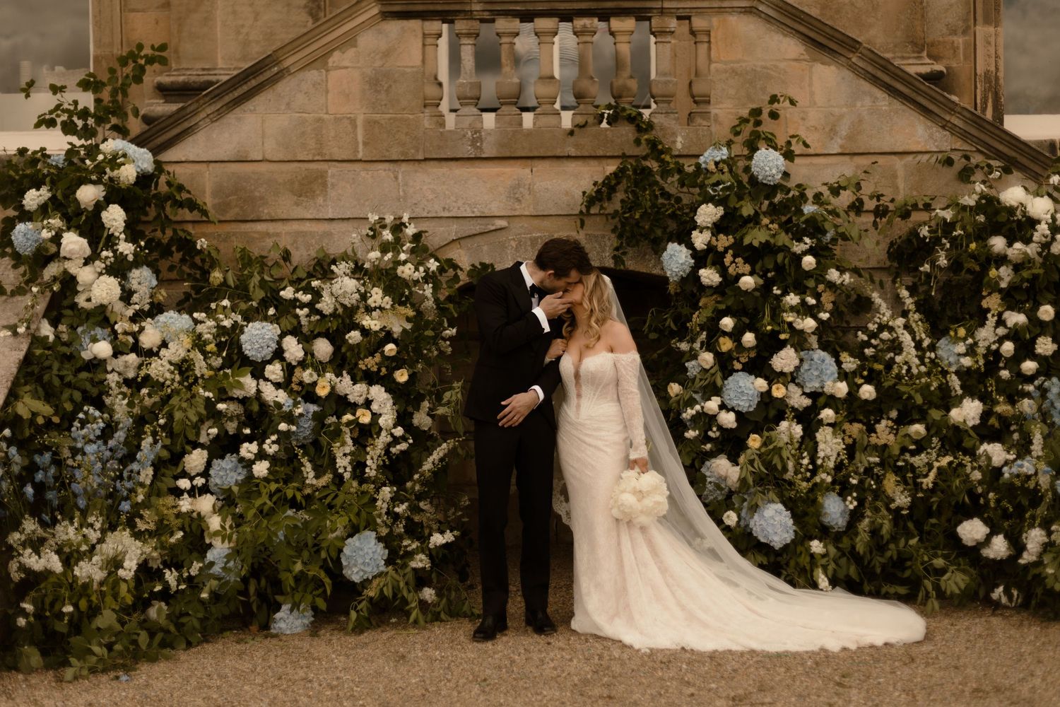 A cheerful wedding party celebrates on the grounds of a historic stone manor house in muted sepia tones.