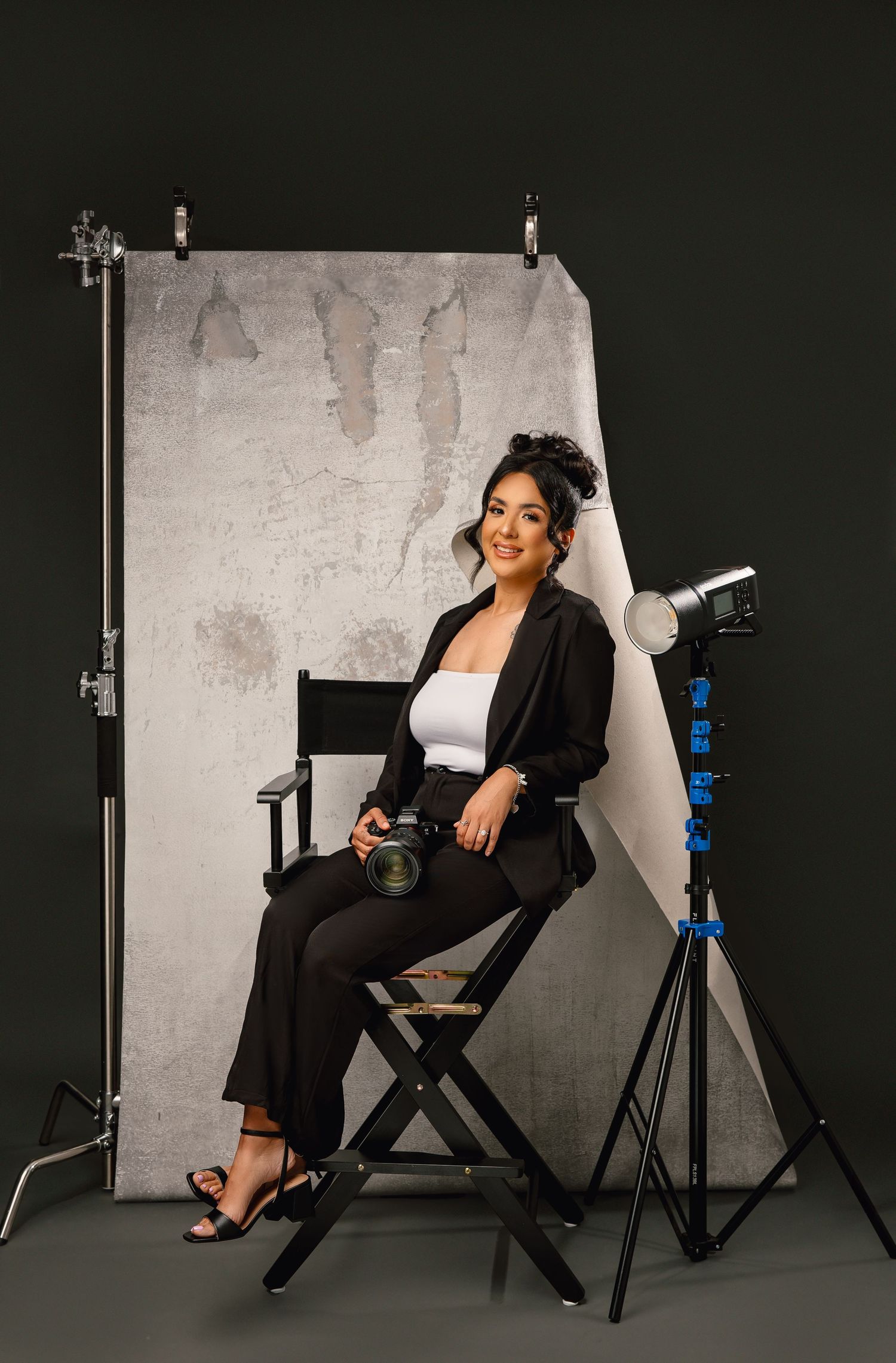 A professional woman in a black blazer and distressed jeans poses confidently while holding makeup brushes against a gray background.