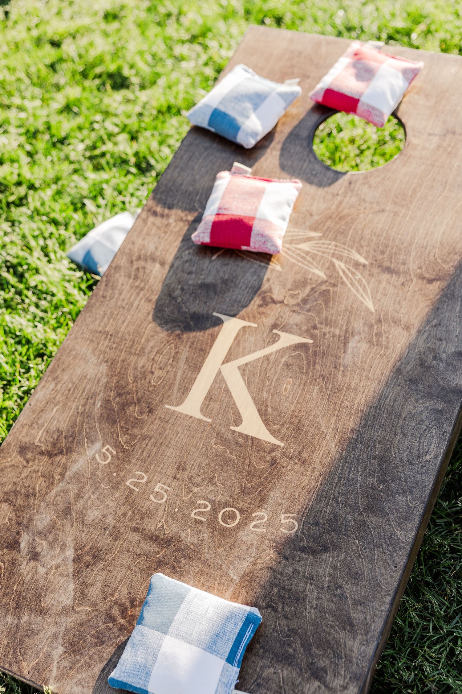 Custom wooden cornhole board with engraved initial 'K' and wedding date displays small bean bags on grass.