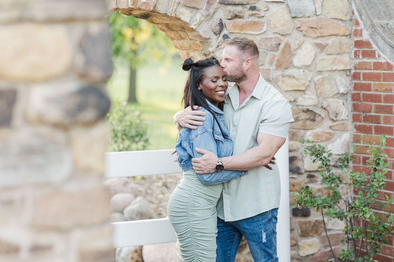 A romantic couple embraces and shares a tender moment against a rustic stone wall during an outdoor photo session.