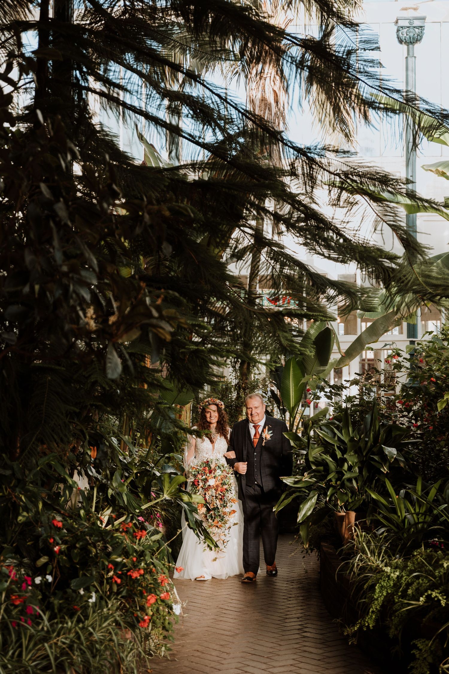 Wedding couple walks along a lush garden path surrounded by tropical plants and red flowers at dusk.