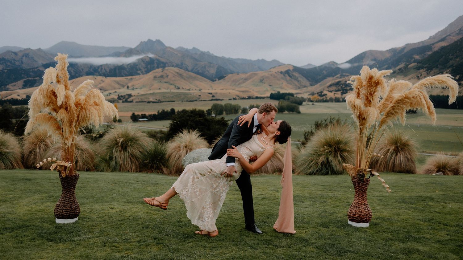A couple dips with pampas grass arrangements in the foreground at a scenic mountain wedding ceremony.