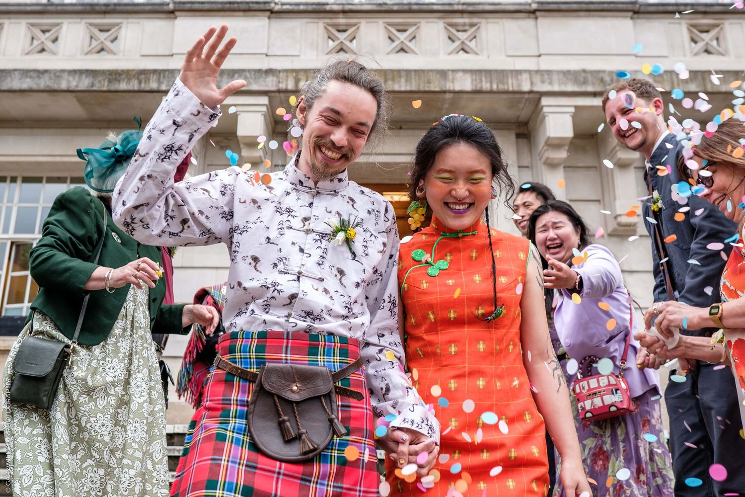 Wedding guests celebrating with confetti outside a historic building as newlyweds smile in traditional Scottish and Asian attire.