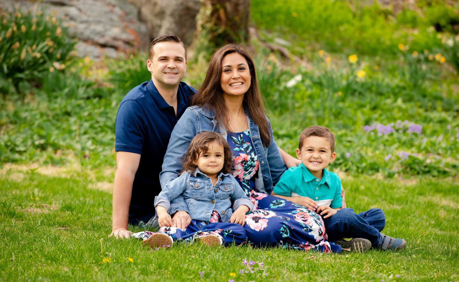 A happy family of four sits together on green grass during an outdoor photo session in a natural setting.