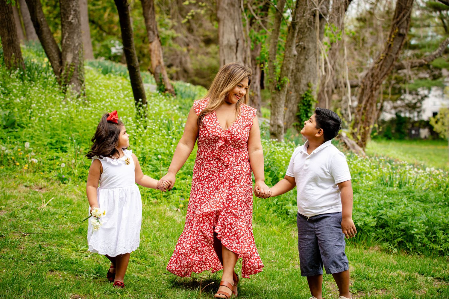 Three people walk hand in hand through a lush green wooded area on a sunny day while wearing casual summer attire.