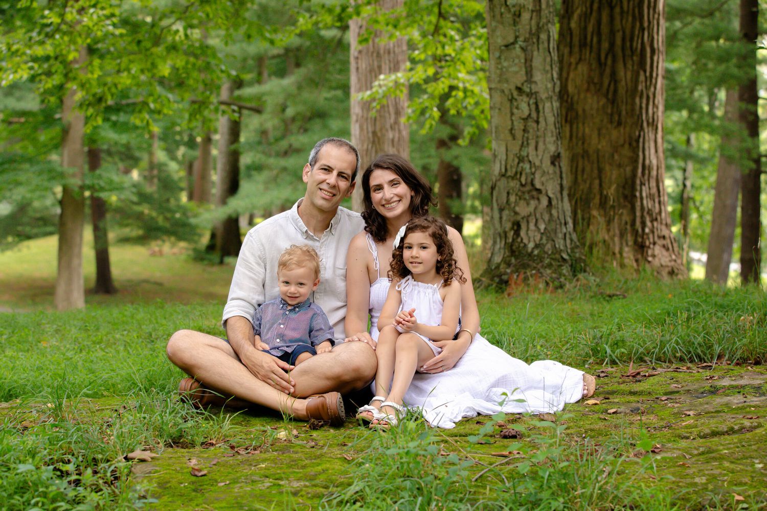 Family relaxing together on grass in lush green forest setting during outdoor photo session.