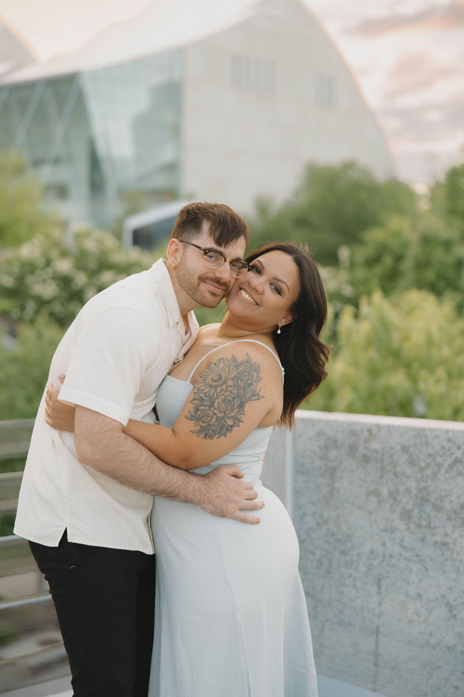 A couple embraces and laughs together outdoors during a romantic photoshoot in front of greenery and modern architecture.