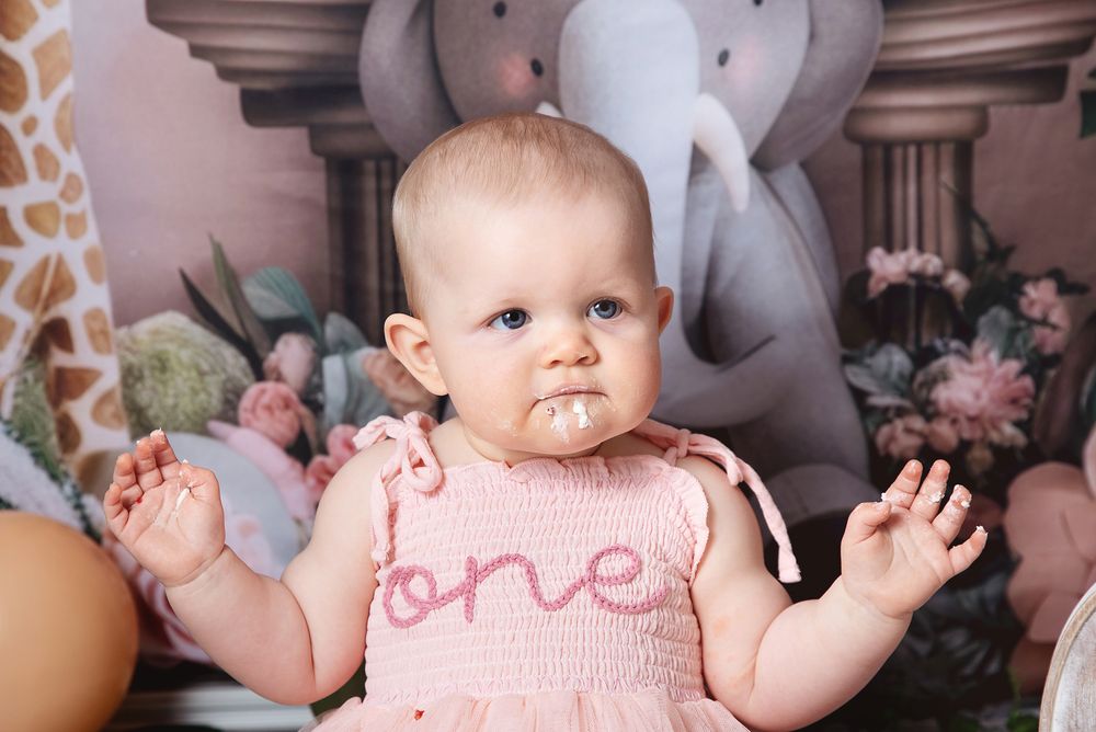 A small baby wearing a pink knit sweater with the word 'one' sits among soft stuffed animals and pillows. During photoshoot in London.
