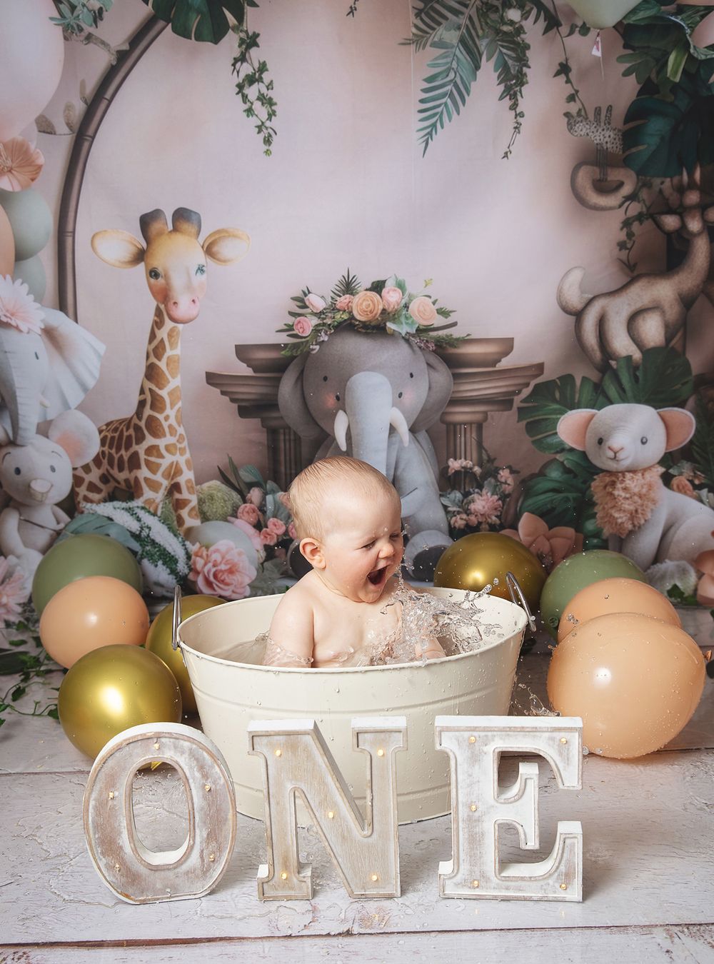 Baby sits in white tub surrounded by safari plush toys, balloons and floral decorations for first birthday photo shoot.