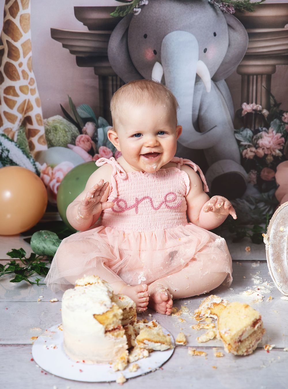 A baby in pink smock dress sits on floor with smashed birthday cake and stuffed elephant decoration in background.
