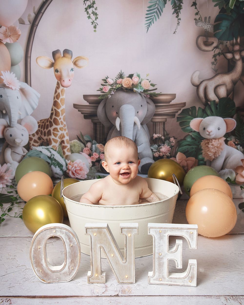 Baby sits in white basin with safari animals, floral decor, gold balloons and ONE white text block for first birthday photo.