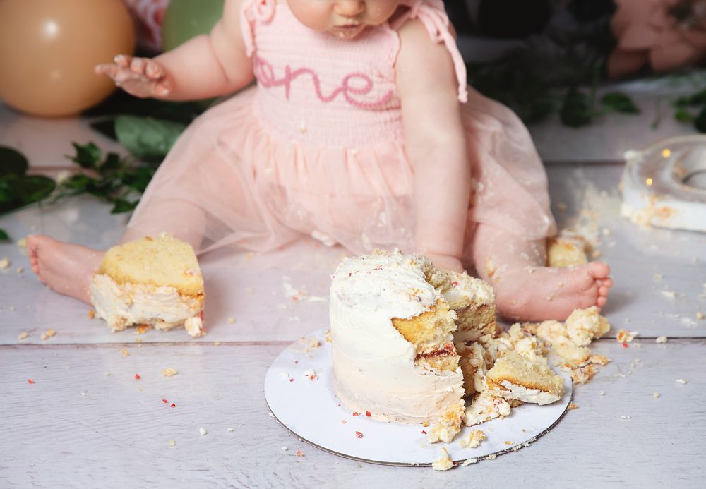 A person dips a finger into frosting during a cake smash photo session with pink and cream tones and scattered decorations.