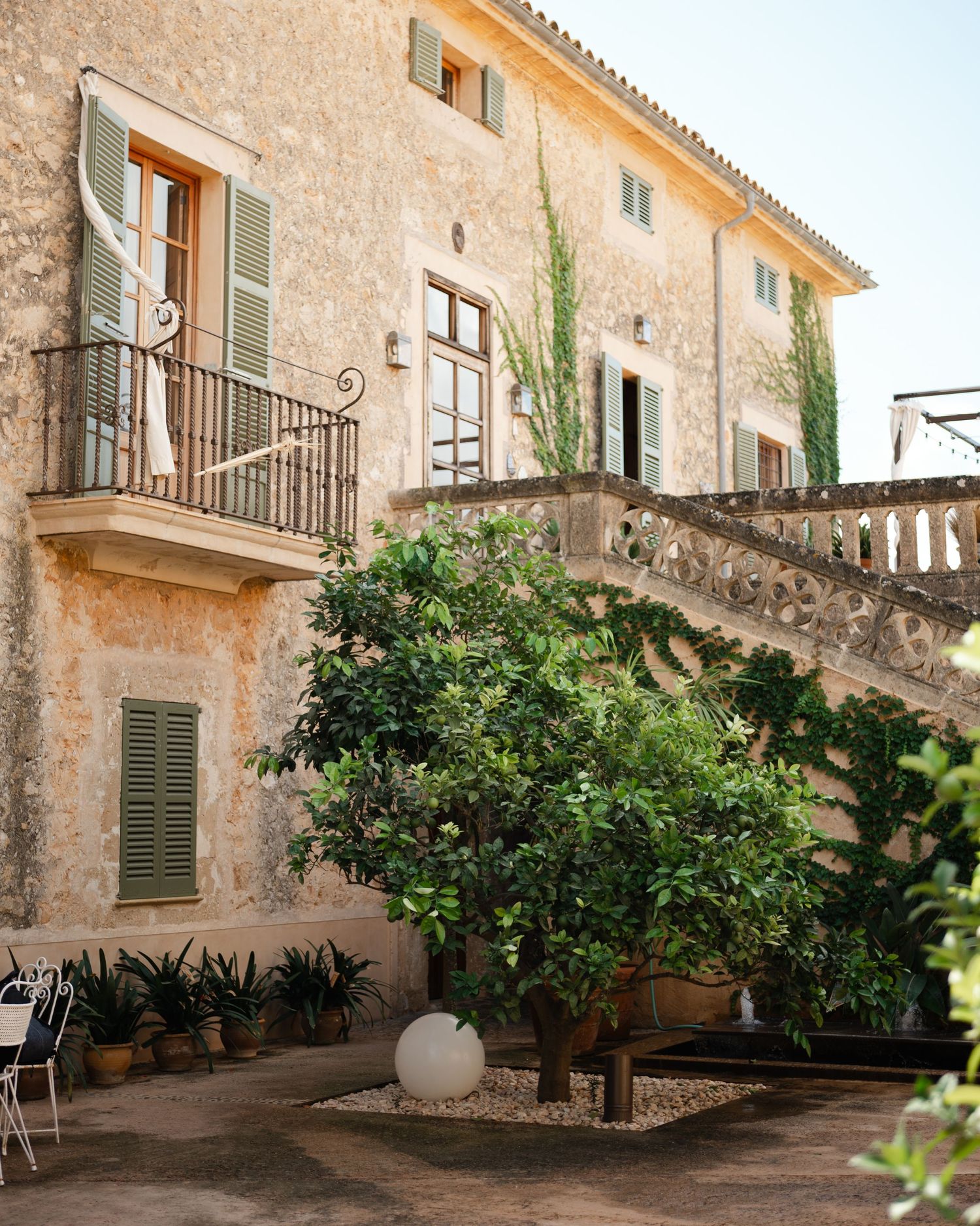 Mediterranean villa with stone facade features a wrought iron balcony, green shutters, and courtyard garden with potted plants.