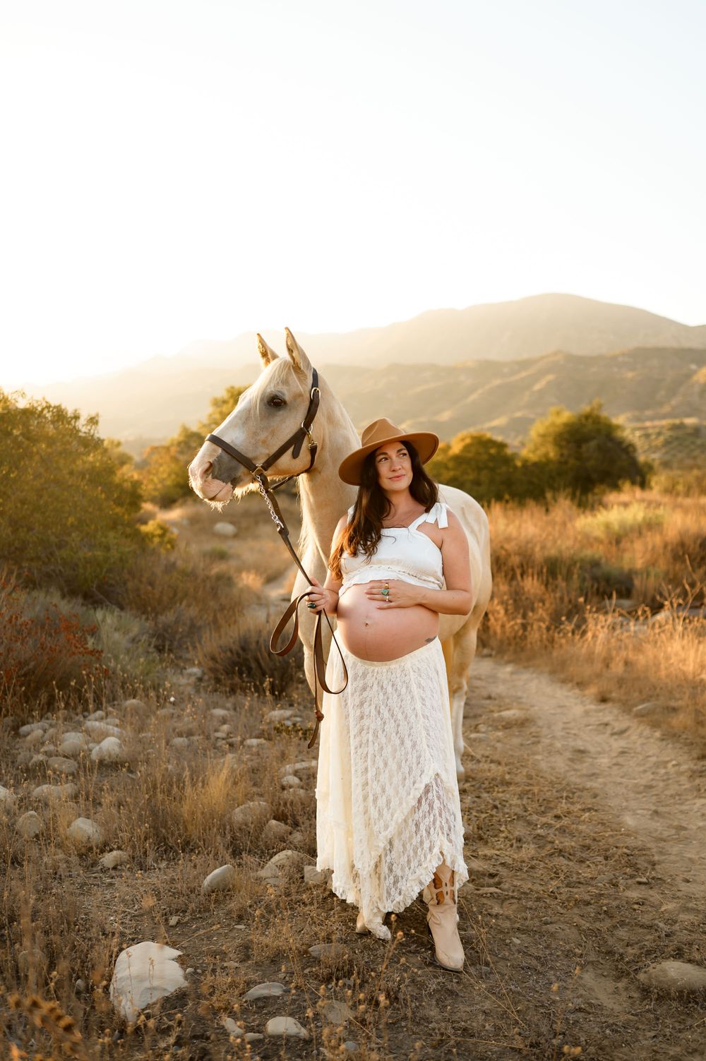 woman stands with white horse in two piece white lace dress at sunset at Ojai Valley Trail riding during her maternity photoshoot in Ojai