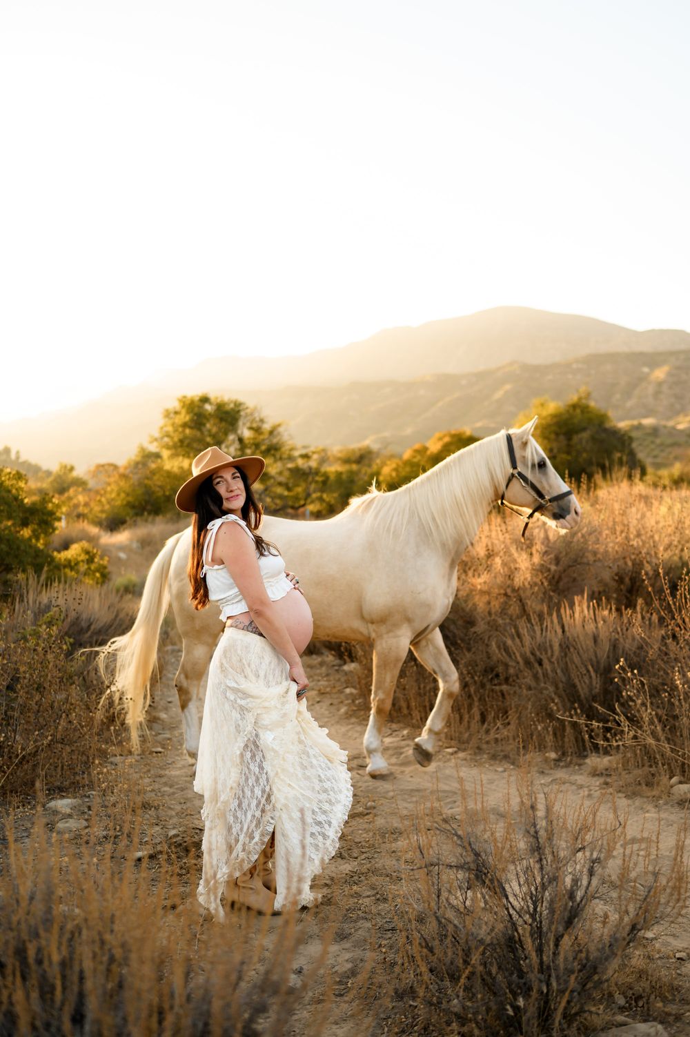 woman poses with white horse in two piece white lace dress and cowboy boots at Ojai Valley Trail riding during her maternity photoshoot