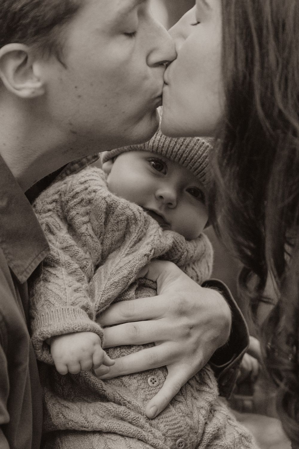 A young child in a knit sweater is embraced between two adults sharing an intimate moment.