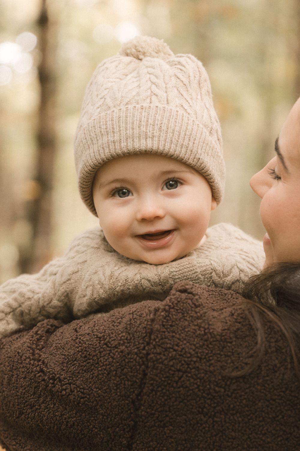 Baby wearing a beige knit beanie and sweater smiles while being held against a blurred autumn background.